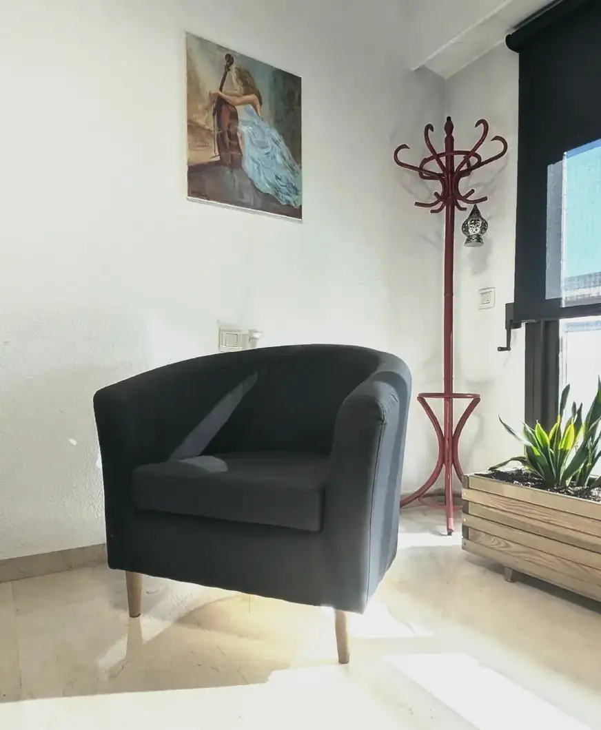 Rincón de la consulta de psicología en Valencia: sillón gris, cuadro, perchero rojo y planta junto a la ventana